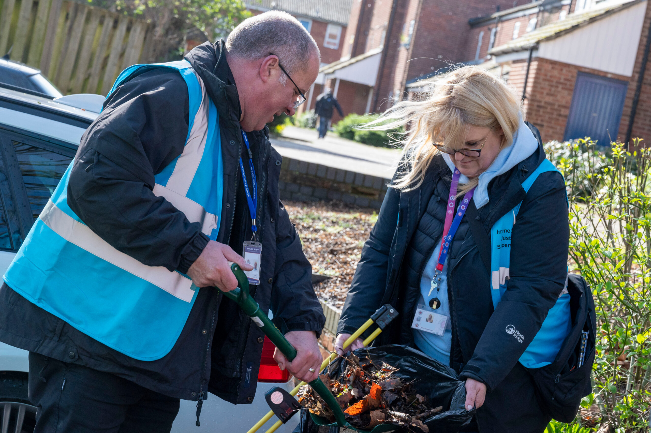 Two people wearing outdoor jackets and high‑visibility vests work together to clear leaves and debris. They are using handheld tools to scoop the material into a black bag. A parked car, fencing, and residential buildings appear in the background on a bright day.