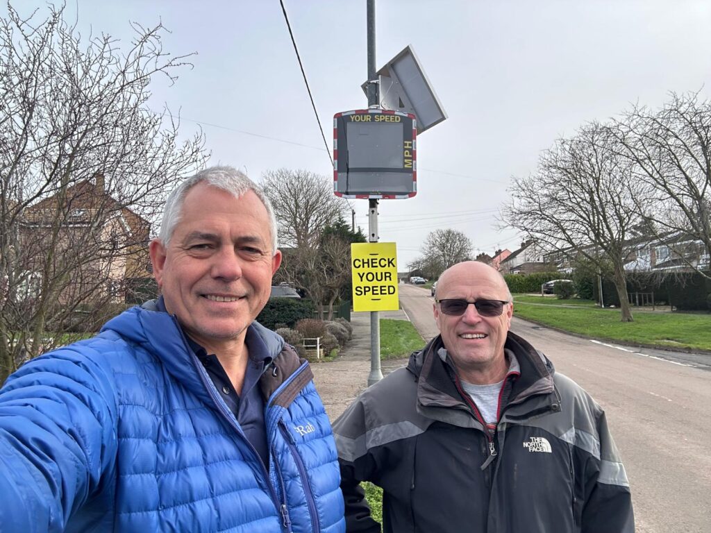 Two people stand on a residential roadside next to a newly installed vehicle-activated speed sign, with a bright yellow “Check Your Speed” sign mounted on the same post. The street is lined with houses, trees, and grass verges under an overcast sky.