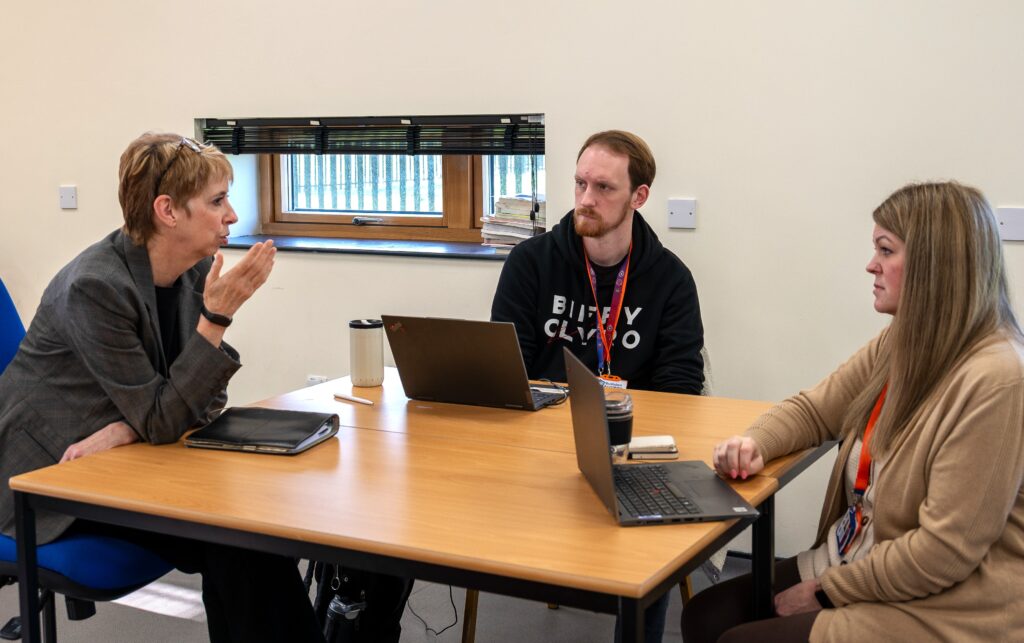 3 people sat indoors around a table, 2 have lap tops