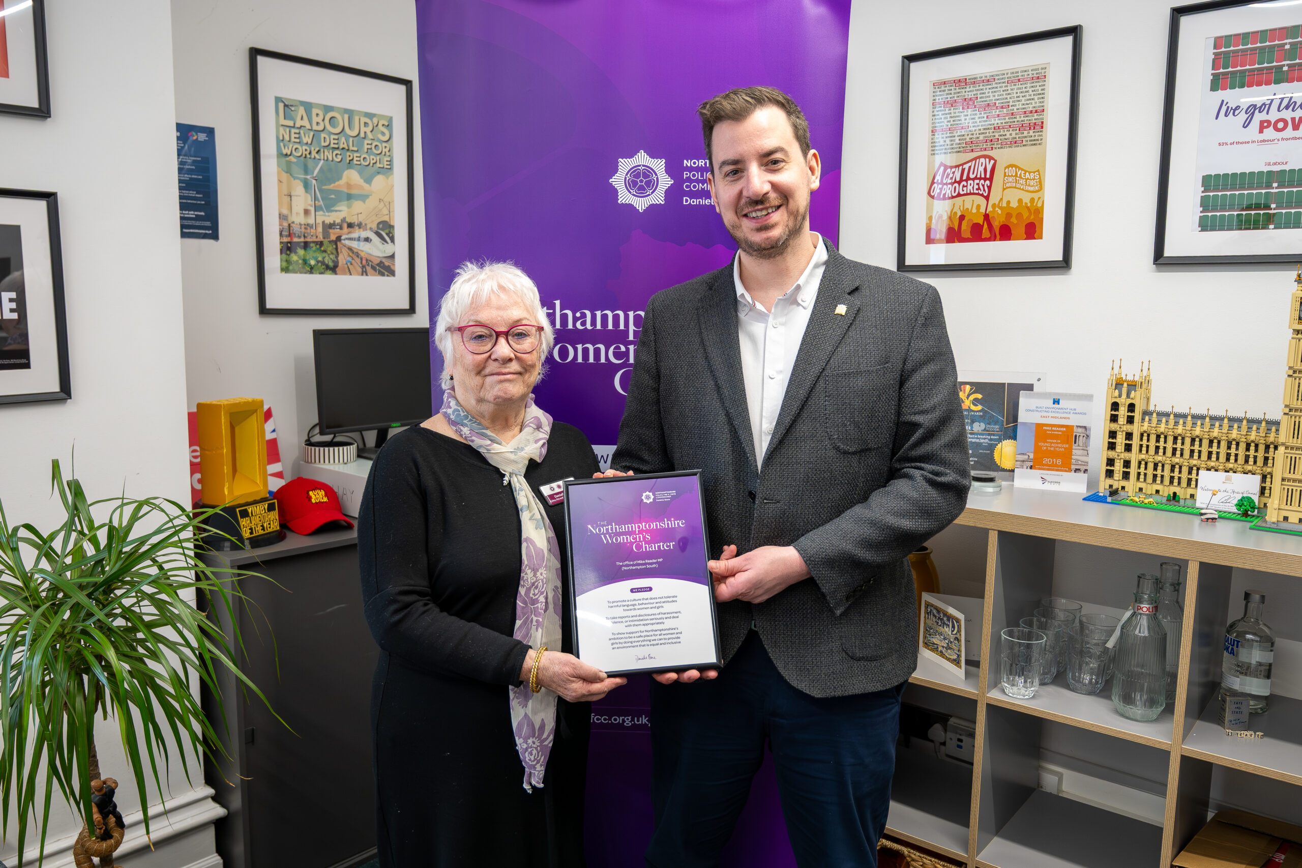 Two people stand indoors holding a framed certificate together. Behind them is a tall purple banner with the Northamptonshire Police, Fire and Crime Commissioner logo. The room has framed posters on the walls, a shelf displaying various model buildings and items, and a potted plant to the left. The certificate is held centrally between the two individuals.
