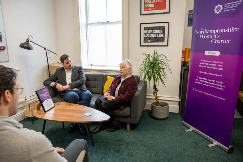 Three people sit in an office-style room having a conversation. Two people sit on a grey sofa with a small wooden coffee table in front of them, where a framed document is displayed. Another person sits opposite them, partially visible in the foreground. A tall purple banner promoting the Northamptonshire Women’s Charter stands to the right, and posters, a potted plant, and a large window are visible in the background.