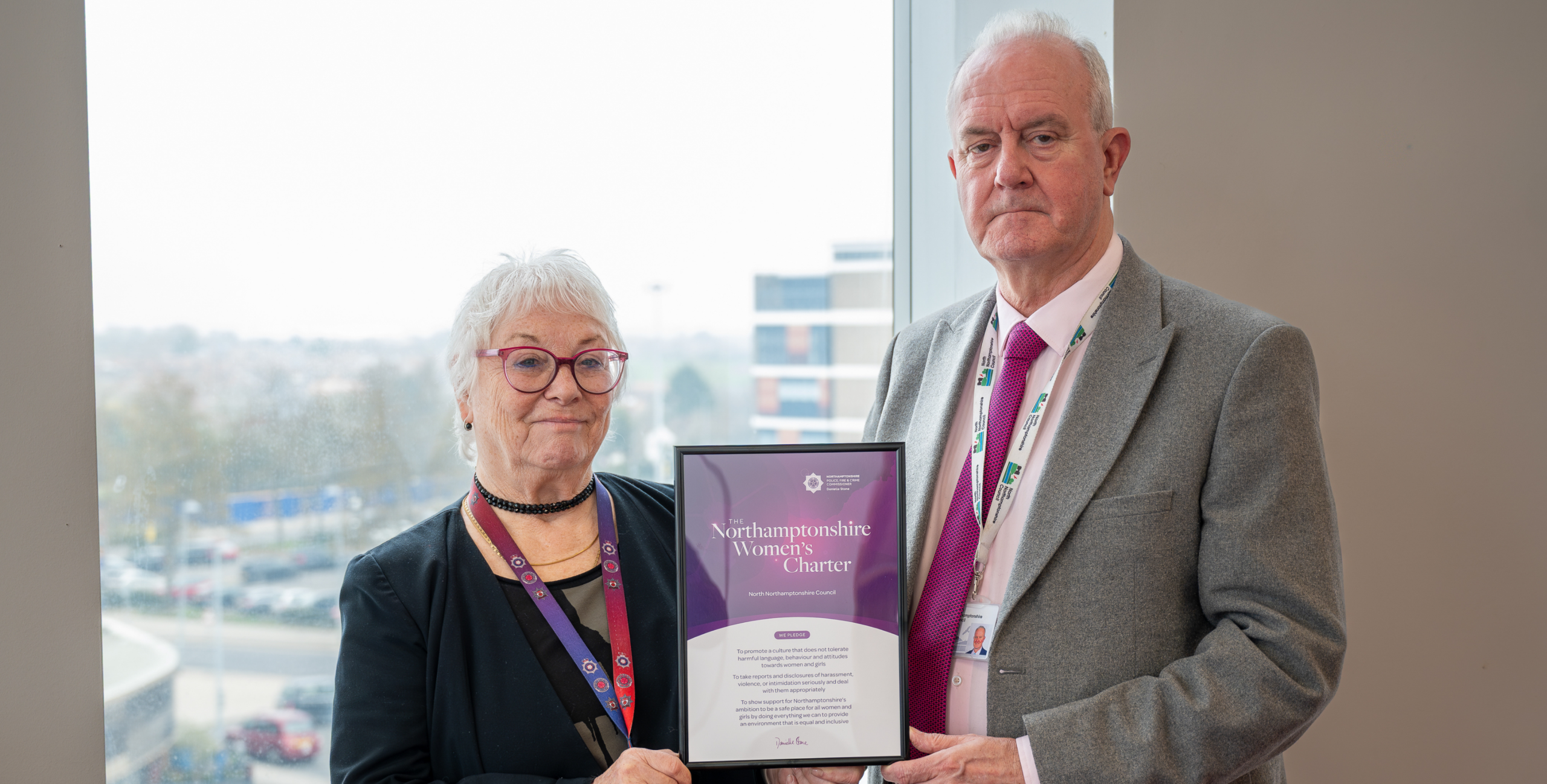Two individuals stand indoors near a large window, holding a framed certificate together. The certificate reads “Northamptonshire Women’s Charter” with additional text beneath it. One individual is dressed in a dark outfit with a lanyard, and the other is wearing a light-colored shirt, a tie, a suit jacket, and a lanyard. A cityscape with buildings and trees is visible through the window in the background.