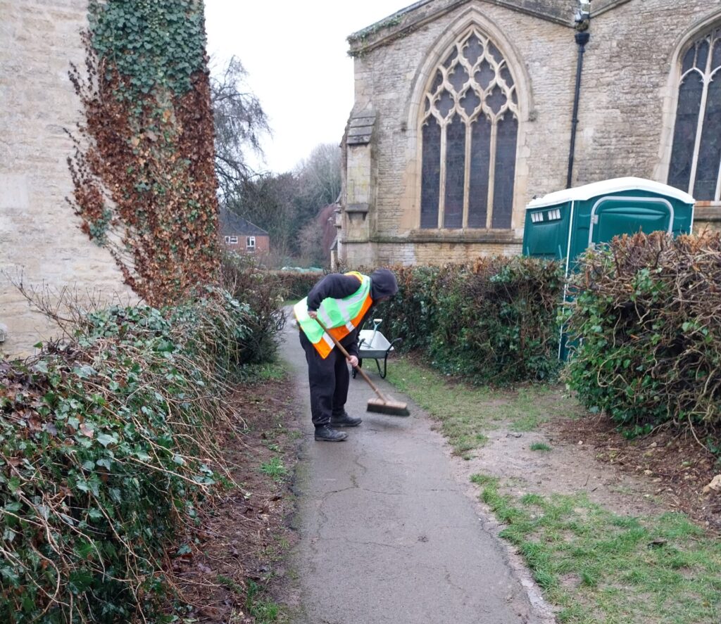 A person wearing a high-visibility vest sweeps a paved path beside a church, bordered by hedges, with a stone church building and a portable toilet visible in the background.