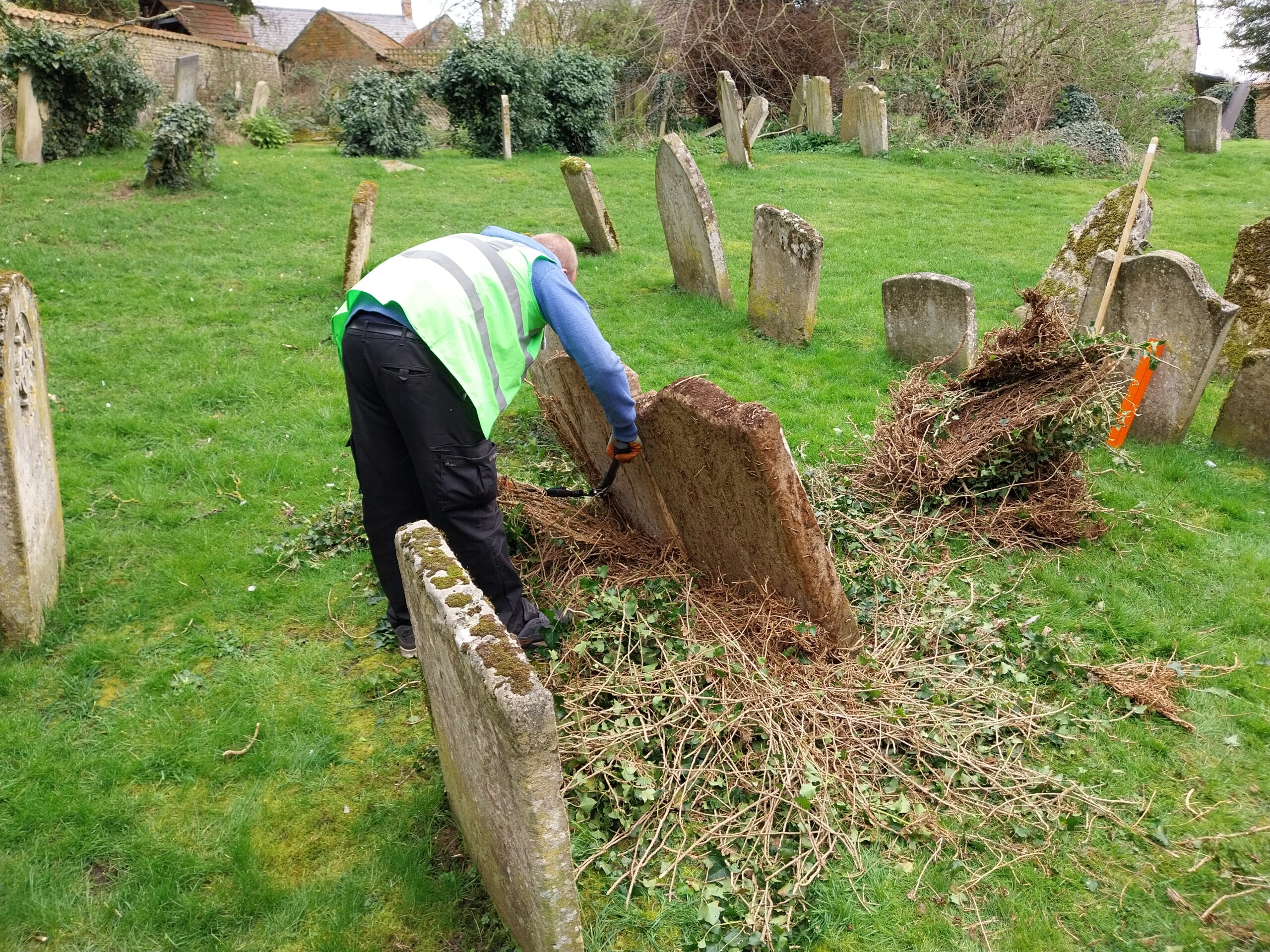 A person wearing a high-visibility vest clears overgrown vegetation around old gravestones in a grassy churchyard, with several weathered headstones standing at different angles in the background.