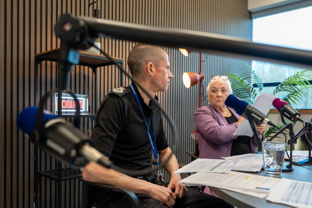 A side view of two people seated at a table in a recording studio. The person on the left wears a dark uniform and faces slightly toward the person on the right, who is wearing a purple blazer and holding papers. Multiple microphones are positioned around them. The background includes a large window with greenery outside and studio lighting fixtures inside. Chief Constable and Commissioner taking part in the second Chat With The Chiefs live broadcast at Darby House on Tuesday, March 31, 2026.