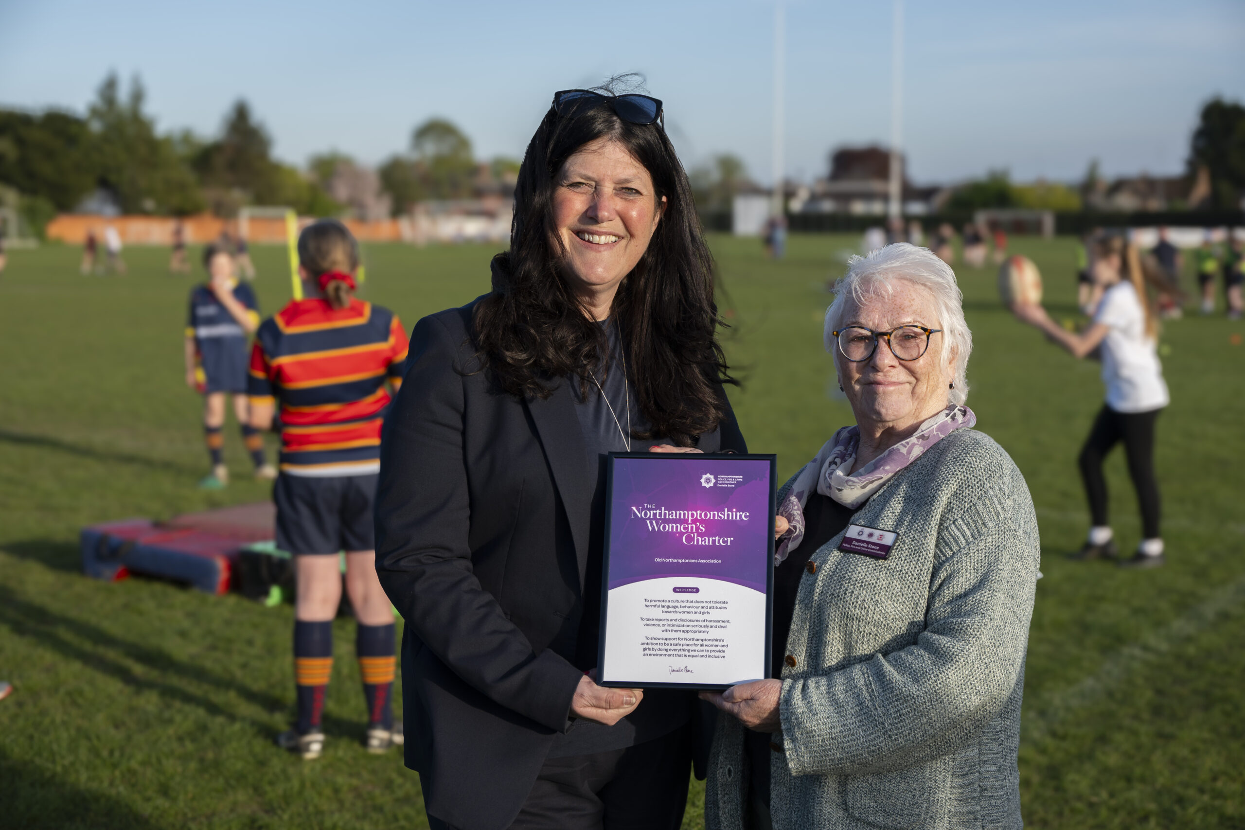 Nikki Clarke-Rogers and Danielle Stone are pictured holding the women's charter certificate infront of rugby players warming up on the pitch.