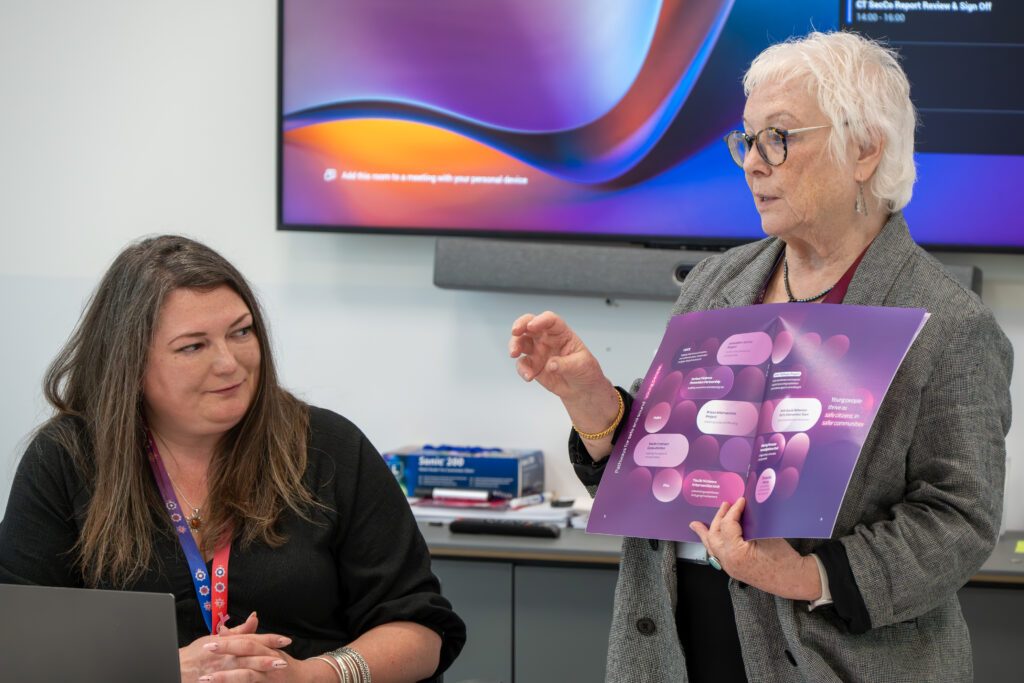 Person standing and speaking while holding a purple presentation booklet during a meeting, with another person seated beside a laptop watching, and a wall-mounted screen in the background.