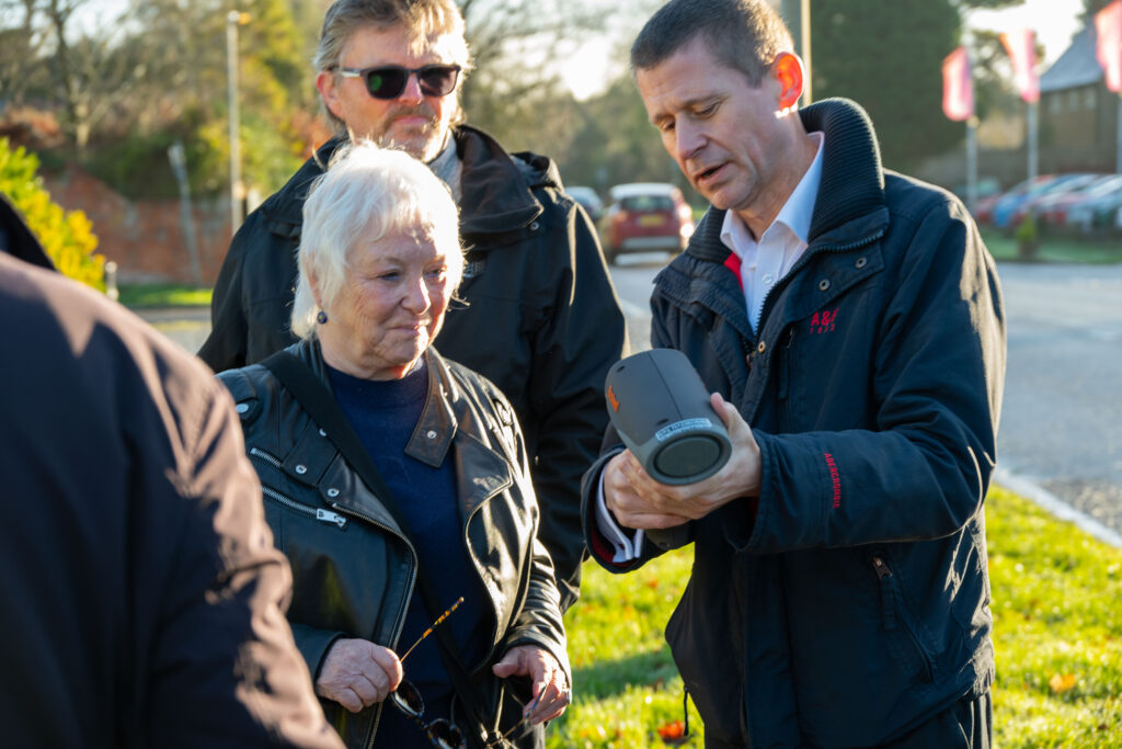 Close-up of several people standing by a roadside as one person holds a handheld speed detection device and shows it to the others, while a residential street with parked cars and greenery appears blurred in the background.