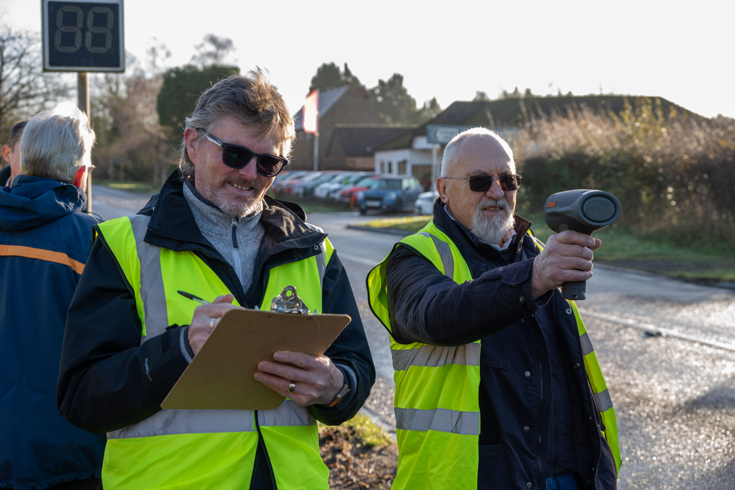 Two people wearing high-visibility vests stand beside a rural roadside measuring vehicle speed, with one person holding a handheld speed gun pointed toward the road while the other records results on a clipboard. In the background, there are parked cars, houses, trees, and a roadside digital speed display showing “88.”