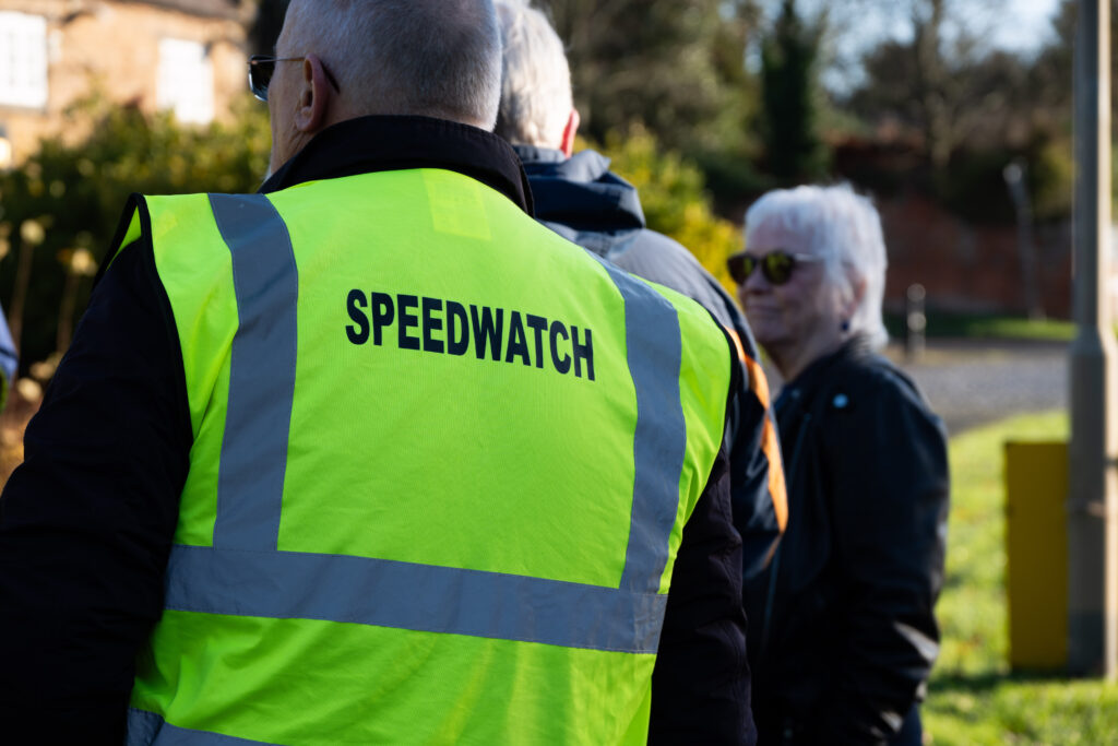 Close-up of a person wearing a high-visibility yellow vest with the word “SPEEDWATCH” printed in large black letters on the back, standing by a roadside. In the background, other people are gathered near greenery and houses, slightly out of focus in bright daylight.