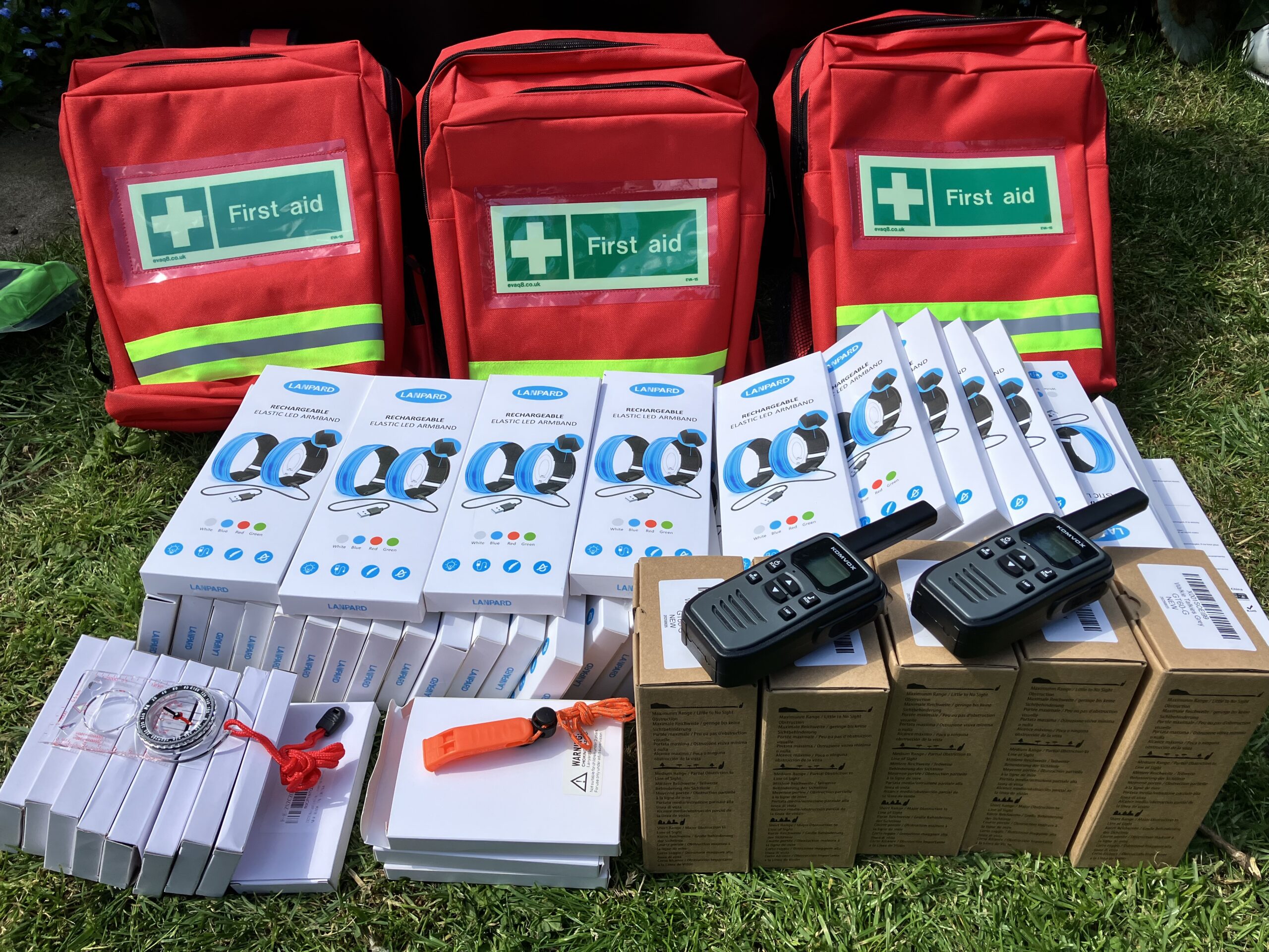 Three bright red first aid backpacks with reflective strips and “First aid” labels are arranged on grass. In front of the bags are neatly stacked emergency items, including boxed rechargeable LED armbands, handheld radios, whistle lanyards, compasses, and other small safety equipment.