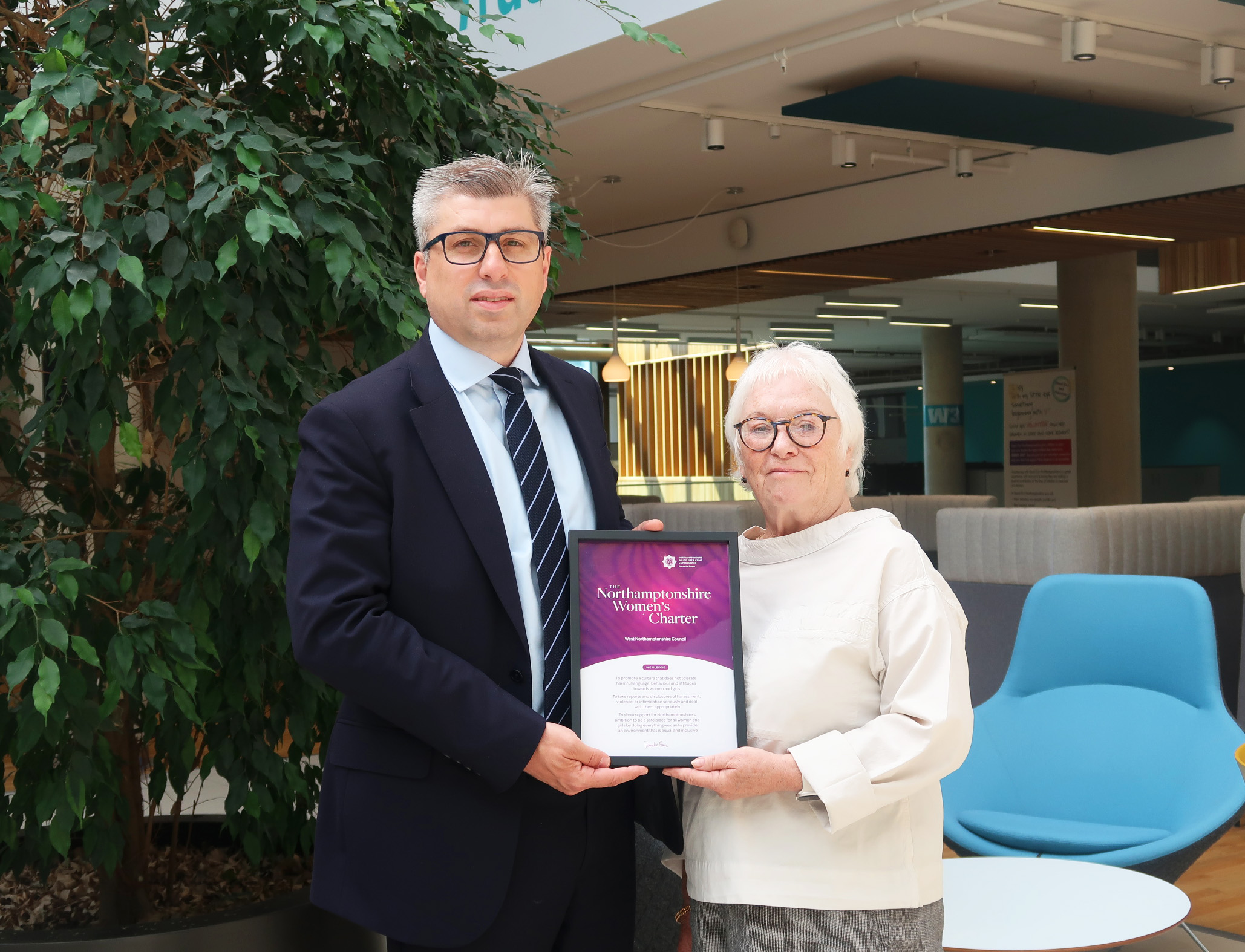Two people in a modern office atrium holding a framed ‘Northamptonshire Women’s Charter’ certificate, with a large indoor plant and seating area in the background