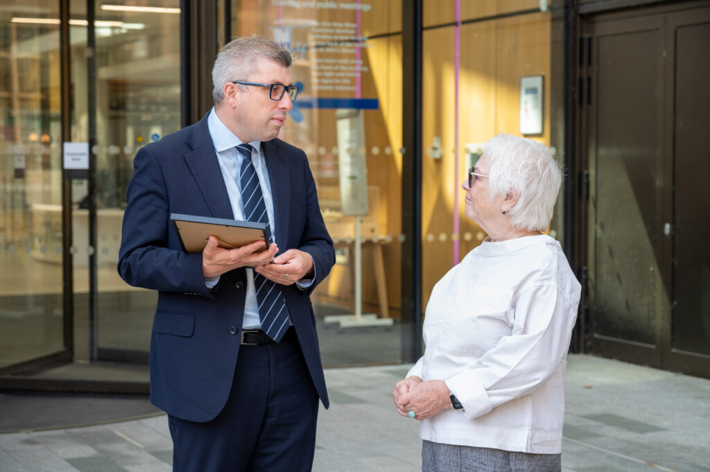 Two people standing outside a glass-fronted building entrance, one holding a framed document while they converse near the doorway