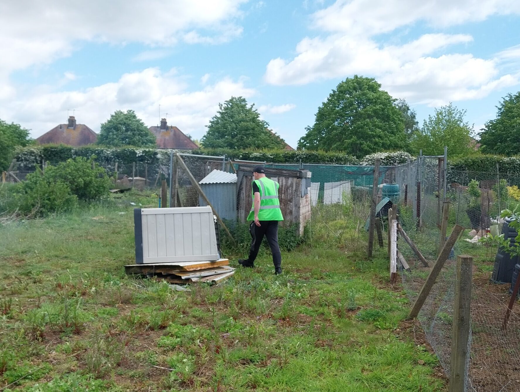 Person wearing a high-visibility vest walking across a grassy allotment site toward a small shed and fenced plots, with scattered weeds, garden structures, and houses and trees in the background under a partly cloudy sky.