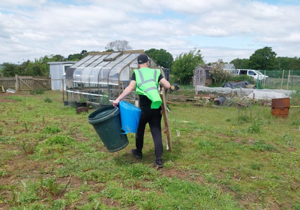 Person wearing a high-visibility vest walking across a grassy allotment site toward a small shed and fenced plots, with scattered weeds, garden structures, and houses and trees in the background under a partly cloudy sky.