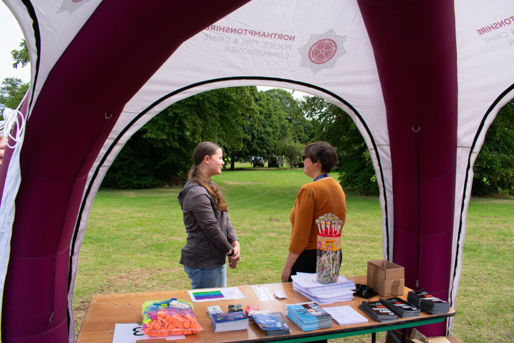 View from inside a maroon and white event tent showing a table with safety leaflets, giveaways, and a container of lollipops in the foreground, while two people stand outside on grass talking, with trees and parked vehicles in the background.