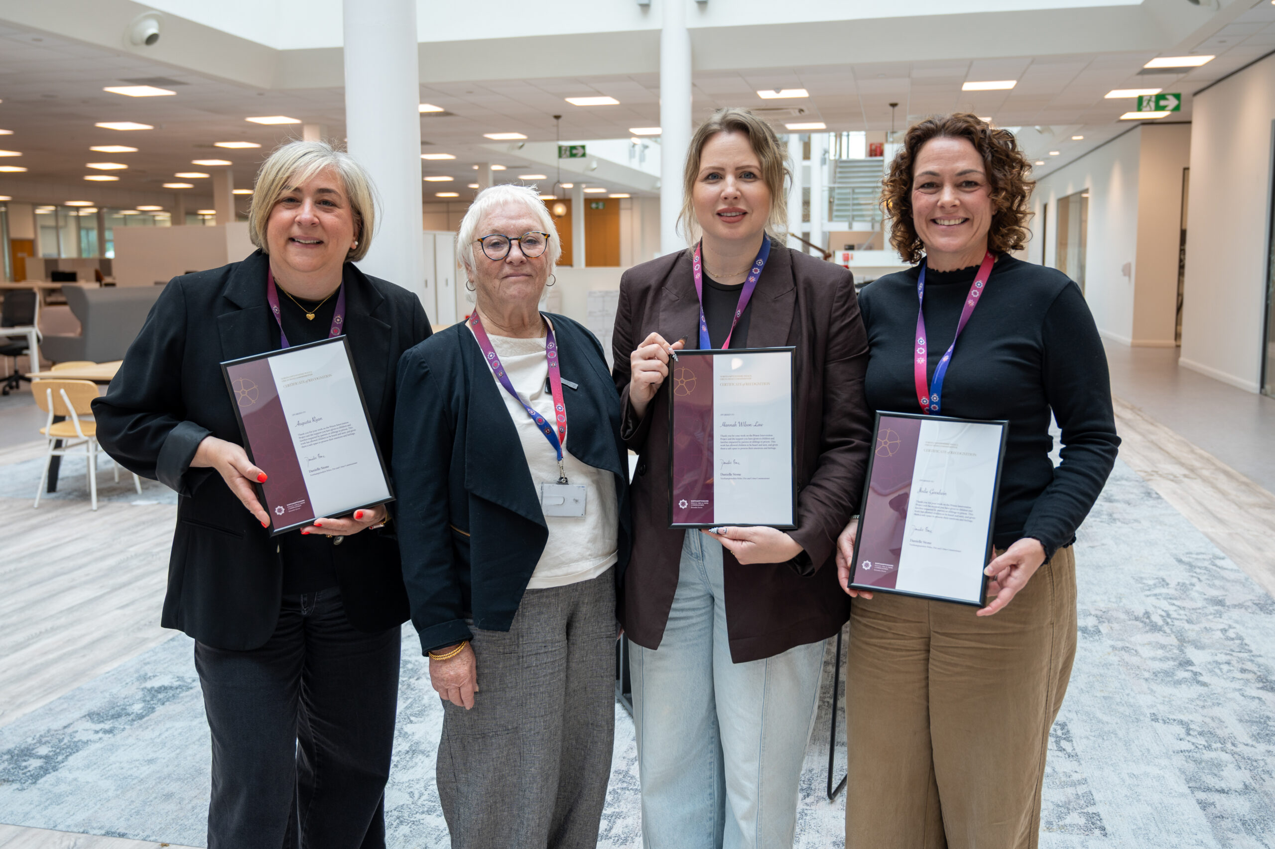 Four people stand in a bright office atrium holding framed certificates of recognition.