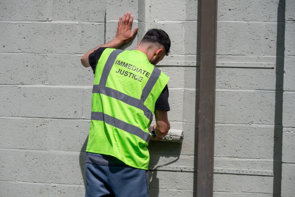 An Immediate Justice participant in a high-visibility vest paints a concrete wall as part of a community payback activity.