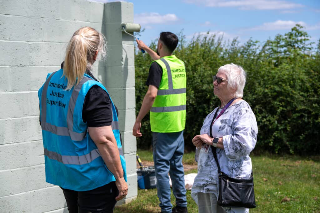 An Immediate Justice participant in a high-visibility vest paints a concrete wall as part of a community payback activity.