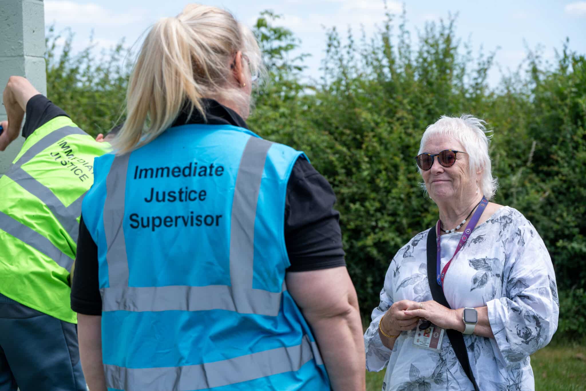 Immediate Justice An Immediate Justice Supervisor wearing a blue high-visibility vest speaks with a community representative outdoors, while an Immediate Justice participant works nearby.