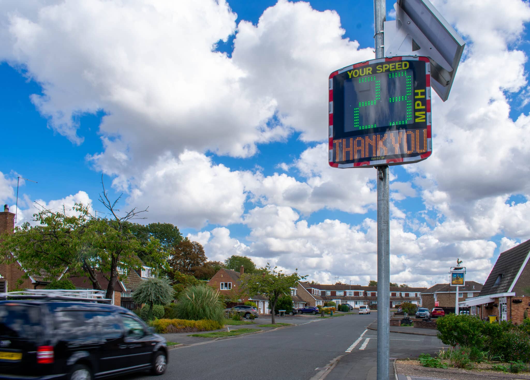 Speed awareness sign on a residential street displaying “24 MPH” and “THANK YOU” beneath a bright, cloudy sky. Houses, trees and parked cars line the road, while a moving black car passes in the foreground.
