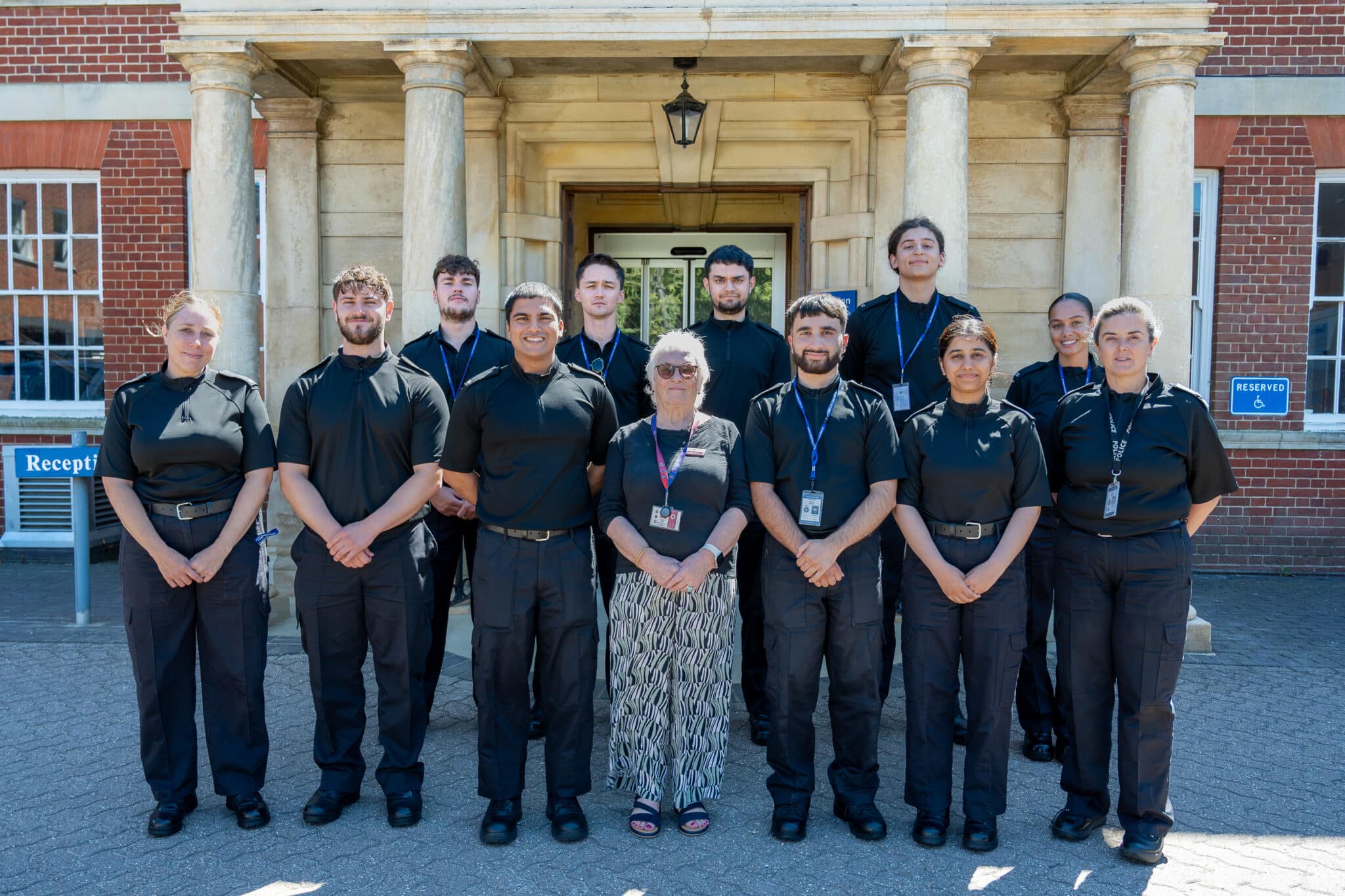Group photograph of 11 people standing outside the entrance of a large brick building with stone columns. Most are wearing dark uniform-style clothing and ID lanyards, with one person standing in the centre wearing a patterned skirt and glasses. The group is arranged in two rows in front of the building's doorway on a sunny day. Visible signs include "Reception" on the left and an accessible parking sign on the right.