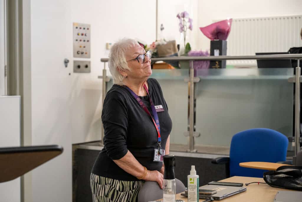 A person with short white hair and glasses stands beside a meeting table in a bright training or conference room. The person is wearing a dark top, patterned skirt and an ID lanyard. On the table are a laptop, mobile phones, a spray bottle and cleaning fluid. In the background, a raised reception-style area with glass panels contains flowers and other items.
