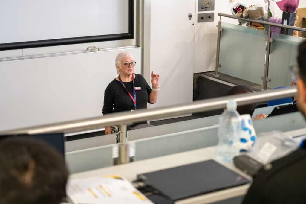 A person wearing glasses and an ID lanyard stands at the front of a tiered training room, speaking to an audience seated out of focus in the foreground. The person gestures while presenting beside a large projection screen. Flowers can be seen on a raised platform behind, and laptops, documents and a water bottle are visible on the desks in front of attendees.