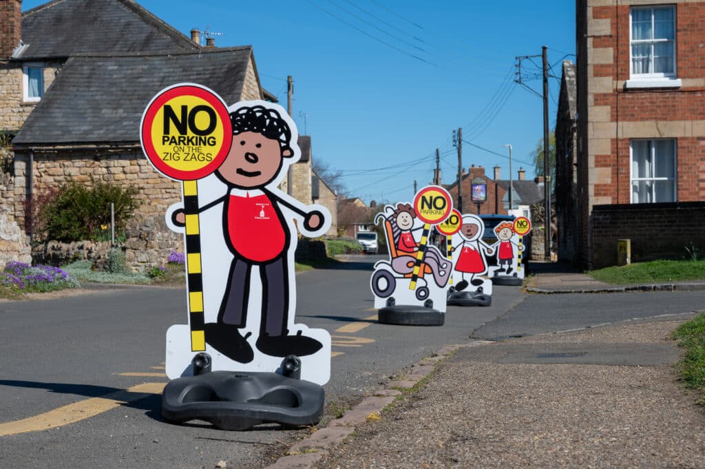 A series of child-shaped road safety signs line a village street outside a school, each holding a circular sign reading “NO PARKING ON THE ZIG ZAGS.” Stone and brick buildings border the road, with the signs positioned along the pavement to remind drivers not to park near the school crossing area.