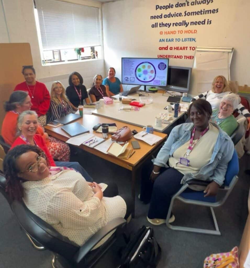 A group of around 12 people are seated and standing around a meeting table in an office training room. Laptops, notebooks, drinks and meeting materials are spread across the table, while a presentation is displayed on a monitor at the front of the room. A large wall quote reads: “People don’t always need advice. Sometimes all they really need is a hand to hold, an ear to listen, and a heart to understand.” A flip chart stands beside the screen, and daylight enters through a window behind the group.