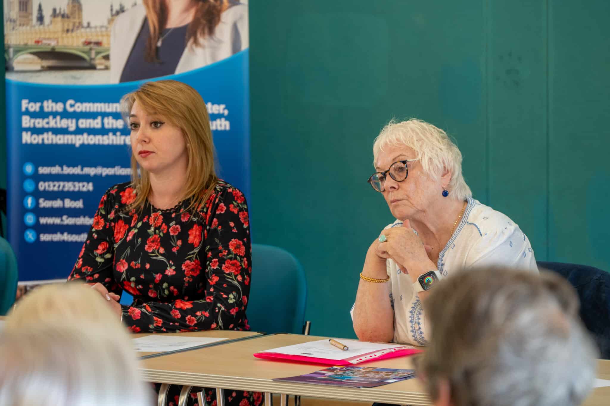 Two people sit at a meeting table during a community discussion. Papers and a pen are on the table in front of them, while attendees are visible in the foreground. A blue community information banner stands behind the group against a teal wall, creating a formal meeting setting.