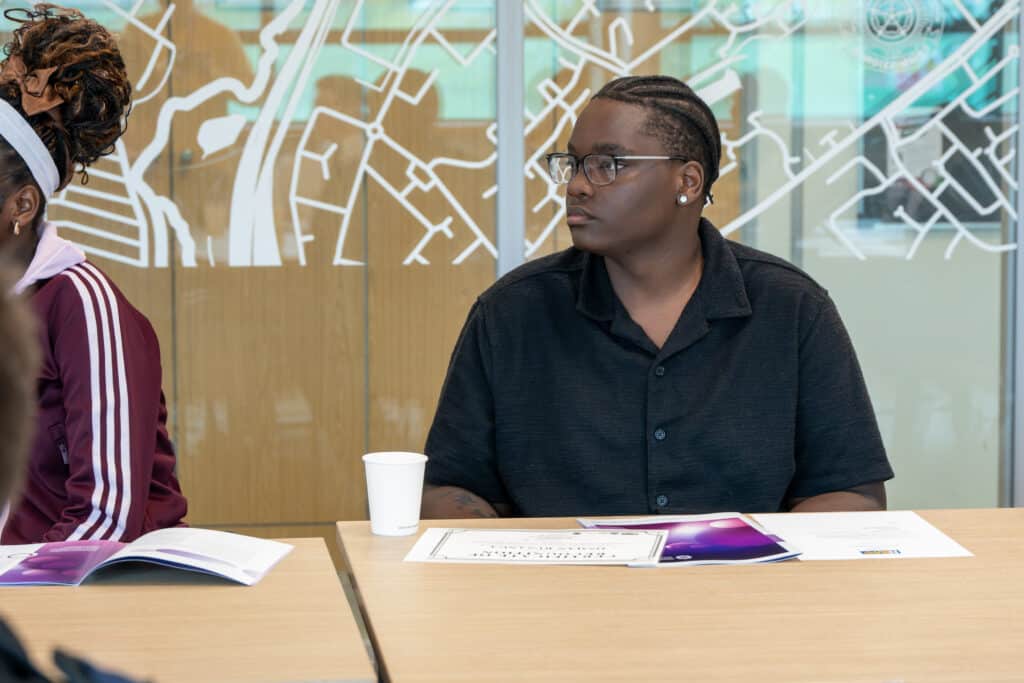 A participant sits at a meeting table listening during a workshop or discussion session. Printed materials and a paper cup are on the table, while a glass wall with a map-style graphic forms the background. Other attendees are partially visible nearby.