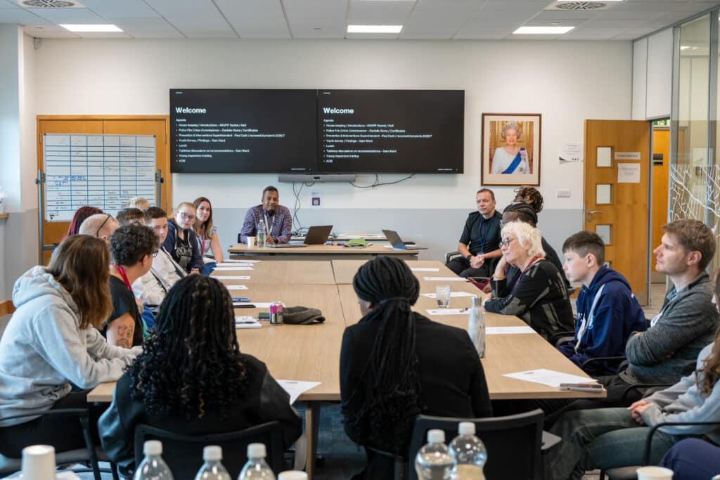 A large group of young people and adults seated around a conference table during a workshop or discussion session. Presentation screens at the front of the room display the meeting agenda, while participants listen and contribute. Papers, laptops and drinks are placed on the table in a modern meeting room.