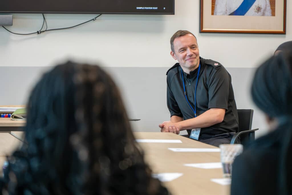 A participant sits at a conference table taking part in a group discussion. Other attendees are visible out of focus in the foreground, while presentation screens and meeting materials can be seen in the room. Papers are laid out on the table during the workshop session.