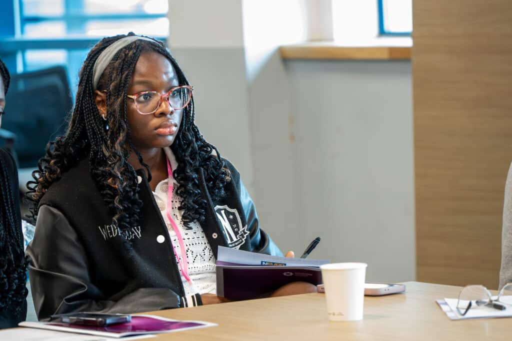 A young participant sits at a meeting table holding a pen and workshop materials while listening during a group discussion. A paper cup, mobile phone and information booklet are on the table, with a bright meeting room visible in the background.